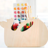 A whale-shaped wooden desk organizer holding paint bottles, paintbrushes, and a watercolor palette, set against a bright, softly blurred background.
