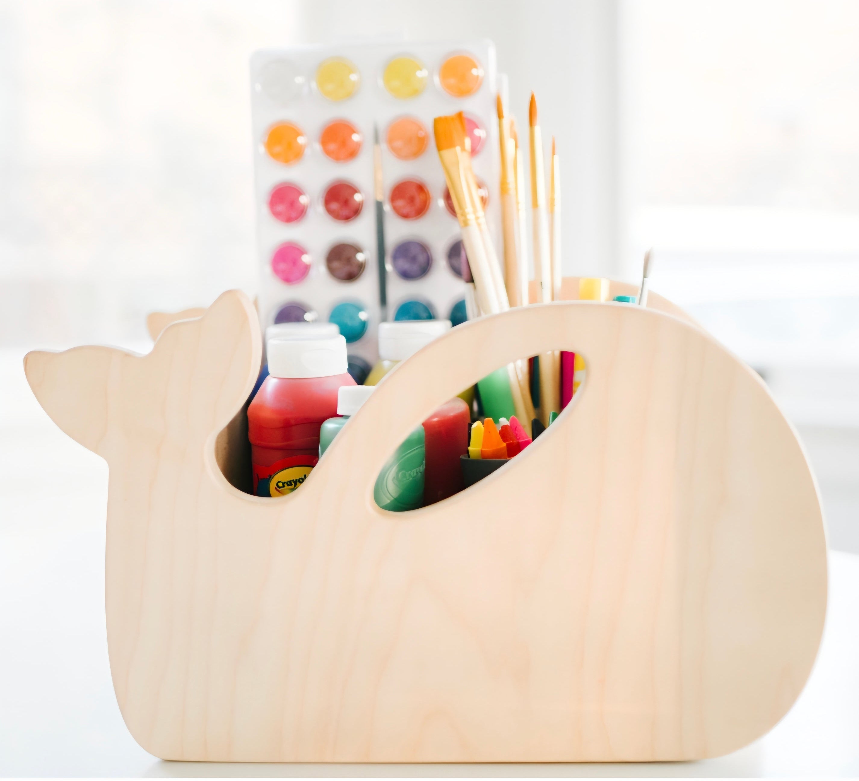 A whale-shaped wooden desk organizer holding paint bottles, paintbrushes, and a watercolor palette, set against a bright, softly blurred background.