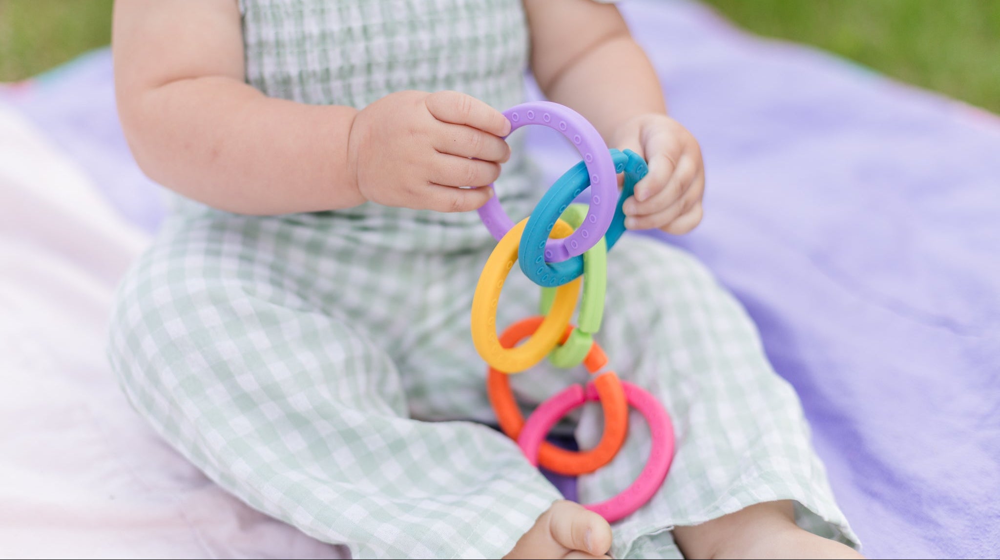 A baby sitting on a purple blanket outdoors, holding a colorful interlocking ring toy.